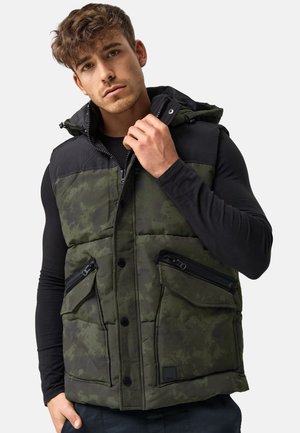 Jeune homme portant un gilet matelassé vert et noir avec fermeture éclair et boutonnage à l'avant, tenant le col d'une main.