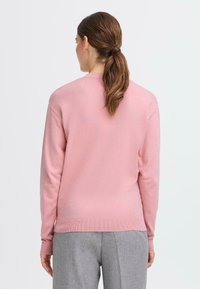 Ljust rosa tröja, stickat tyg, rund hals, ribbade mudd och nederkant, bärs med grå byxor, visad bakifrån.