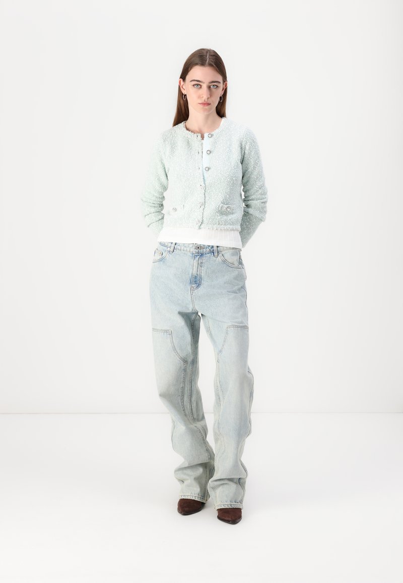 Hellgrüner strukturierter Cardigan mit rundem Ausschnitt und silbernen Knöpfen, kombiniert mit lockeren hellblauen Jeans und braunen spitz zulaufenden Schuhen.