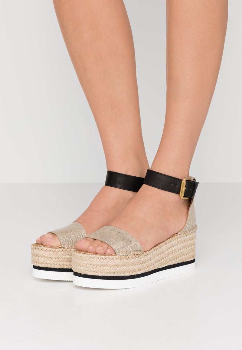 Sandales compensées espadrilles beiges avec brides noires à la cheville et boucles dorées, portées pieds nus sur un fond neutre.