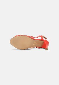 Une sandale rouge à talon haut montrée de la semelle, avec un talon bas, une bride à la cheville et une semelle extérieure beige texturée.