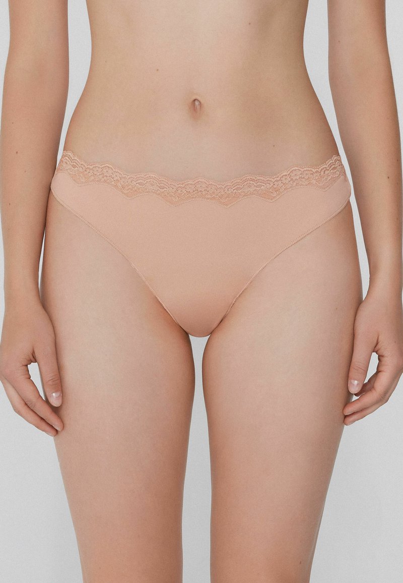 Beige Gefäß mit Spitzenbesatz aus einem glatten, weichen Stoff. Verfügt über einen gewellten Spitzenbund, der für Komfort und eine nahtlose Passform sorgt.