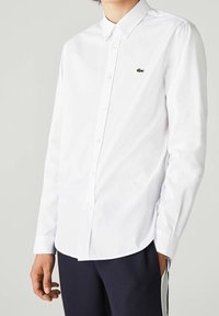 White long-sleeve shirt in cotton with button-up design, pointed collar, and small green embroidered logo on the chest.
