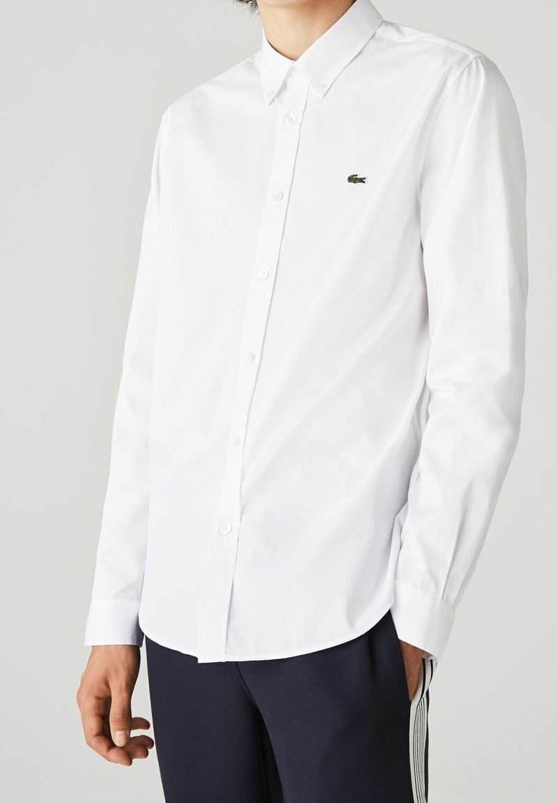 White long-sleeve shirt in cotton with button-up design, pointed collar, and small green embroidered logo on the chest.