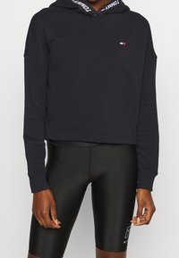 Sudadera corta de color azul marino con mangas largas, que cuenta con una capucha ajustable y acentos de logo. Combinada con pantalones cortos negros ajustados.