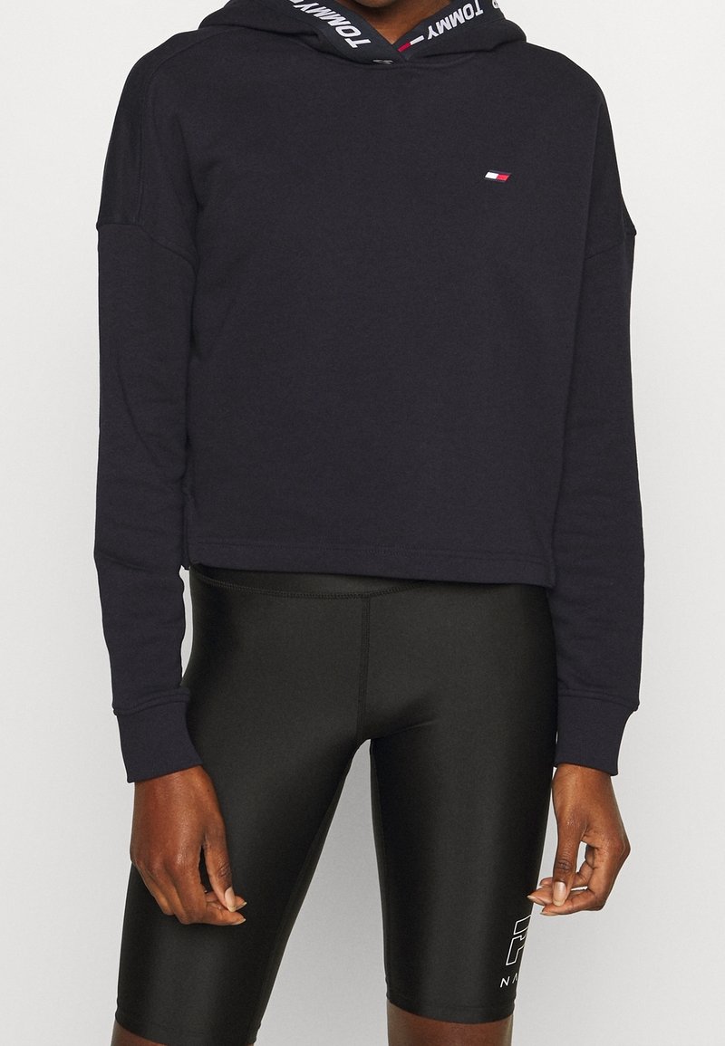 Sudadera corta de color azul marino con mangas largas, que cuenta con una capucha ajustable y acentos de logo. Combinada con pantalones cortos negros ajustados.