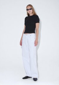 Top ajustado de manga corta negro combinado con jeans anchos blancos. La modelo lleva gafas de sol negras y zapatos de punta negros tipo slip-on.