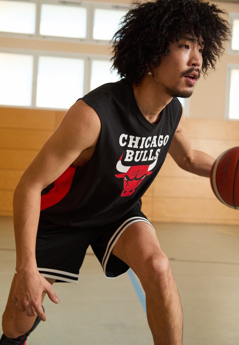 Melnā bezpiedurkņu basketbola krekls ar "CHICAGO BULLS" tekstu un sarkaniem akcentiem, kombinēts ar melnajiem šortiem, kas apmīti ar baltām svītrām. Basketbols rokā.