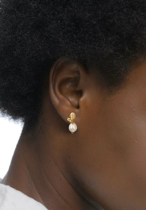 Goldene Ohrringe mit einem Blätterdesign und einer tropfenförmigen weißen Perle. Glatte Textur mit metallic glänzendem Finish, getragen auf einem dunklen Hautton.
