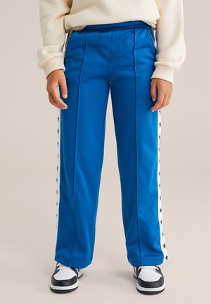 Enfant portant un pantalon de survêtement bleu avec des panneaux latéraux rayés blancs étoilés et des baskets noir et blanc, les mains détendues sur les côtés.