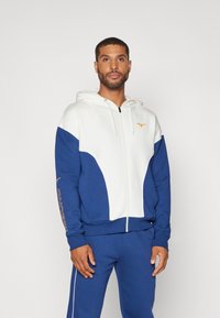 Mikina s kapucňou na zips, ktorá má biele a tmavomodré panely, s rebrovanými manžetami a lemom, a s oranžovým logom na hrudi a rukáve.