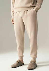 Pantaloni de trening bej dintr-un material texturat, cu talie elastică și buzunare laterale, asortați cu pantofi slip-on din suede gri, cu catarame.