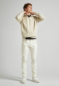 Sweatshirt beige à col zippé sur une chemise noire, associé à un jean blanc et des baskets blanches. Le tissu a une texture douce avec un minimum de branding.