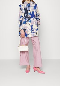 Nainen pukeutuneena pastelliväriseen abstraktikuosisateenkaaritakkiin, vaaleanpunaisiin leveälahkeisiin housuihin, vaaleanpunaisiin korkokenkiin, pitelee pientä valkoista käsilaukkua, jossa on kultainen lukko.