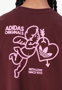Kastanjebrun bomulds T-shirt med et pink grafik af en engel, der holder et hjerte med Adidas-logoet. Teksten siger "ADIDAS ORIGINALS" og "WITH LOVE SINCE 1972."