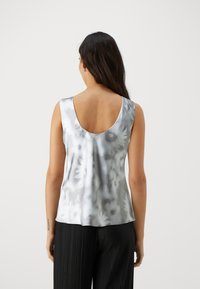 Silberne Satin-Top ohne Ärmel mit einem runden Rückenausschnitt, das ein florales Design in verschiedenen Grautönen zeigt. Kombiniert mit schwarzen, plissierten Hosen.