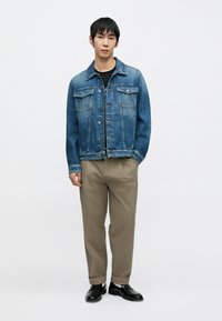 Veste en denim bleu moyen avec poches poitrine et boutons argentés, associée à un pantalon beige et des chaussures noires, debout devant un fond uni.
