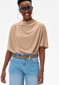 Uśmiechnięta kobieta w dużych okularach przeciwsłonecznych w oprawkach typu żółw, dużych kolczykach kołach, beżowej bluzce z drapowaniem, niebieskich dżinsach i brązowym pasku ze złotą klamrą.