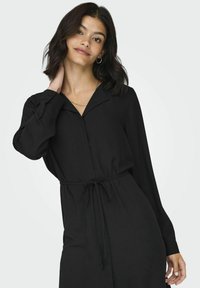 Abito lungo nero con maniche a lunghe e colletto, vita regolabile con coulisse e texture liscia. Sottopunti dettagli con bottoni sulle maniche.