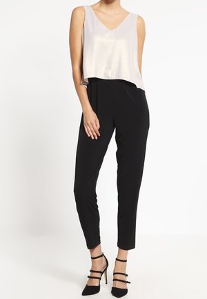 Vrouw draagt mouwloze losse lichtbeige top, aansluitende zwarte broek en zwarte hoge hakken met enkelbandjes, staand tegen een witte achtergrond.