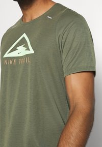 Olivgrön kortärmad Nike Trail-tröja med en strukturerad väv, prydlig med en ljusgrön triangel-logotyp och beige text nedanför.