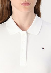 Valge polo särk tekstuuriga kangast, millel on krae ja kuus nuppu, ning vasakul rinnal väike logo punaste ja siniste aktsentidega.