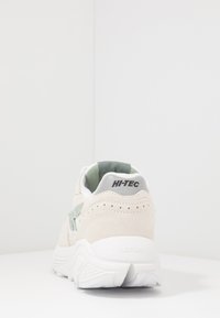 Beige suede sneaker with green accents and a white sole. Features a padded collar and perforated detailing above the heel.