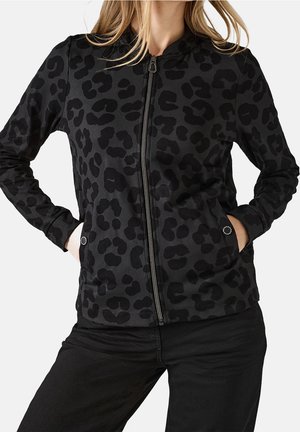 Kvinde iført en sort lynlåstrøje med mørkt leopardprint, hænderne i forlommerne, kombineret med sorte bukser.