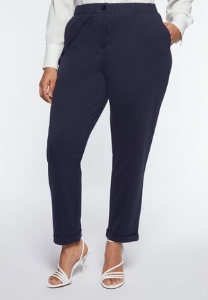 Pantalon bleu marine ajusté avec taille élastique, ourlets retroussés, porté avec des sandales blanches à talons hauts et lanières, et un haut blanc à manches longues.