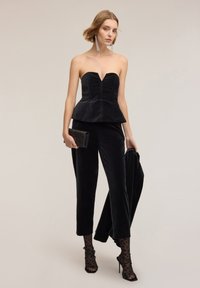 Kobieta ubrana w czarny, bezramiączkowy top z basenką, dopasowane spodnie, przezroczyste rajstopy w panterkę, czarne szpilki, trzymająca dopasowaną kurtkę i błyszczącą kopertówkę.