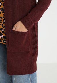 Burgundy lång cardigan med ribbat mönster, framfickor och avslappnad passform, över en djärv djurmönstrad topp och jeans.