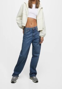 Veste courte matelassée crème, crop top blanc, jean large bleu et baskets grises. Détails remarquables : poignets élastiques sur la veste, style denim classique.