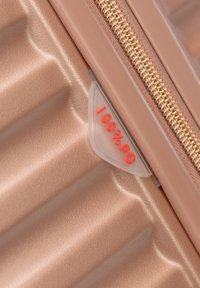Primo piano di un angolo di una valigia in oro rosa con texture, dotata di una cerniera in bronzo e un'etichetta trasparente che mostra "100%PC" in rosso.
