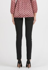 Svarta figurnära byxor i kombination med en blus med geometriskt mönster i mörkröd och rosa ton; svarta högklackade skor fulländar outfiten.