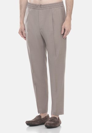 Uomo che indossa pantaloni su misura marrone chiaro con vita elasticizzata e mocassini in pelle scamosciata marrone scuro, in piedi contro uno sfondo semplice.