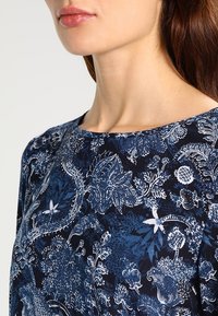 Marineblaues Oberteil mit floralem Muster, das weiße botanische Designs zeigt, runder Ausschnitt und glatte Stofftextur. Details an den Ärmelrändern sichtbar.