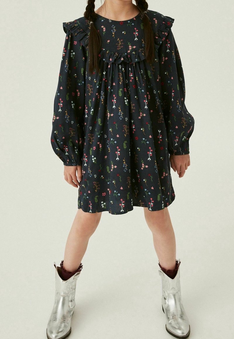 Enfant dans une robe fleurie sombre avec des volants et des manches longues, portant des bottes de cowboy argentées brillantes et des chaussettes bordeaux.