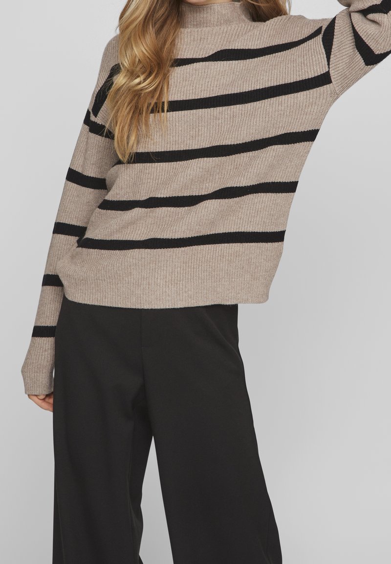 Femme portant un pull côtelé beige avec des rayures horizontales noires et un pantalon noir ample sur un fond clair uni.