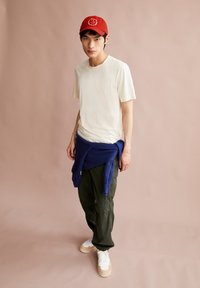 Weißes Baumwoll-T-Shirt, marineblauer Pullover um die Taille gebunden, olivgrüne Cargo-Hose und beige-weiße Sneakers. Rote Kappe mit Logo.