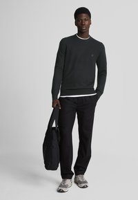 Suéter verde oscuro con cuello redondo, con un ribete blanco en contraste, combinado con pantalones negros y un bolso negro de hombro. Zapatillas deportivas con detalles en gris.