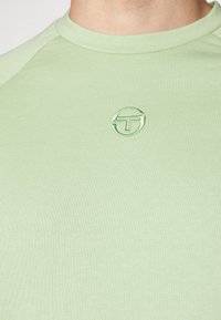 T-shirt côtelé vert clair à col rond avec un petit logo rond brodé représentant un T stylisé sur la poitrine.