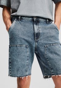 Shorts en denim bleu clair avec une finition délavée, présentant des bords effilochés, de grandes poches latérales et un design classique à cinq poches.