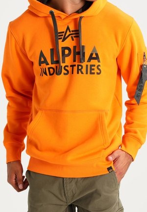 Man draagt een feloranje Alpha Industries hoodie met zwart logo, zwarte trekkoorden, voorzak en groene cargobroek.