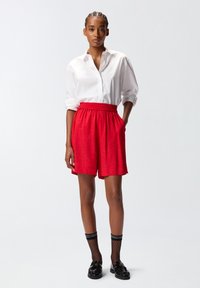 Pantaloni corti rossi con cintura elastica, abbinati a una camicia bianca con bottoni. Calze nere a rete e mocassini completano l'outfit.