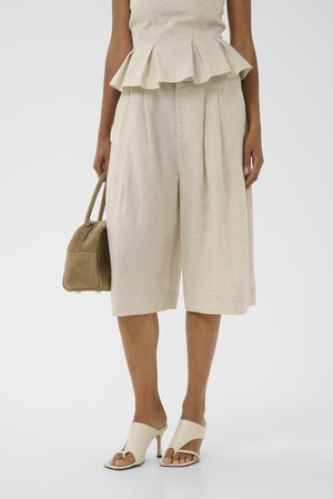Vrouw draagt beige culotte shorts met wijde pijpen en bijpassende peplum top, houdt een beige handtas vast en draagt crèmekleurige mules met hoge hak.