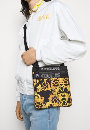 Schwarze Schultertasche mit kunstvollem goldenen Muster, mit Reißverschluss versehenen Fächern und einem strukturierten Stoff. Das Versace Jeans Couture-Logo ist sichtbar platziert.