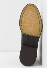 Semelle de chaussure présentant un avant-texturé beige, un milieu en bois foncé avec couture, et un talon foncé lisse, posée contre un mur d'angle blanc.
