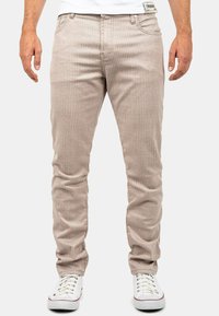 Pantalons en velours côtelé beige avec des rayures verticales, design slim fit, cinq poches, et une étiquette blanche à la taille. Associés avec des baskets blanches.
