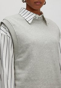 Grauer ärmelloser Pullover mit gerippten Kanten über einem weißen Hemd mit blauen vertikalen Streifen und einem spitzen Kragen.