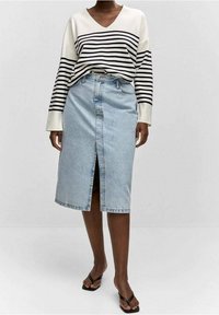 Femme portant un haut rayé à manches longues avec col en V, une jupe en jean bleu clair longueur genou avec une fente à l'avant, et des sandales noires ouvertes à l'avant.